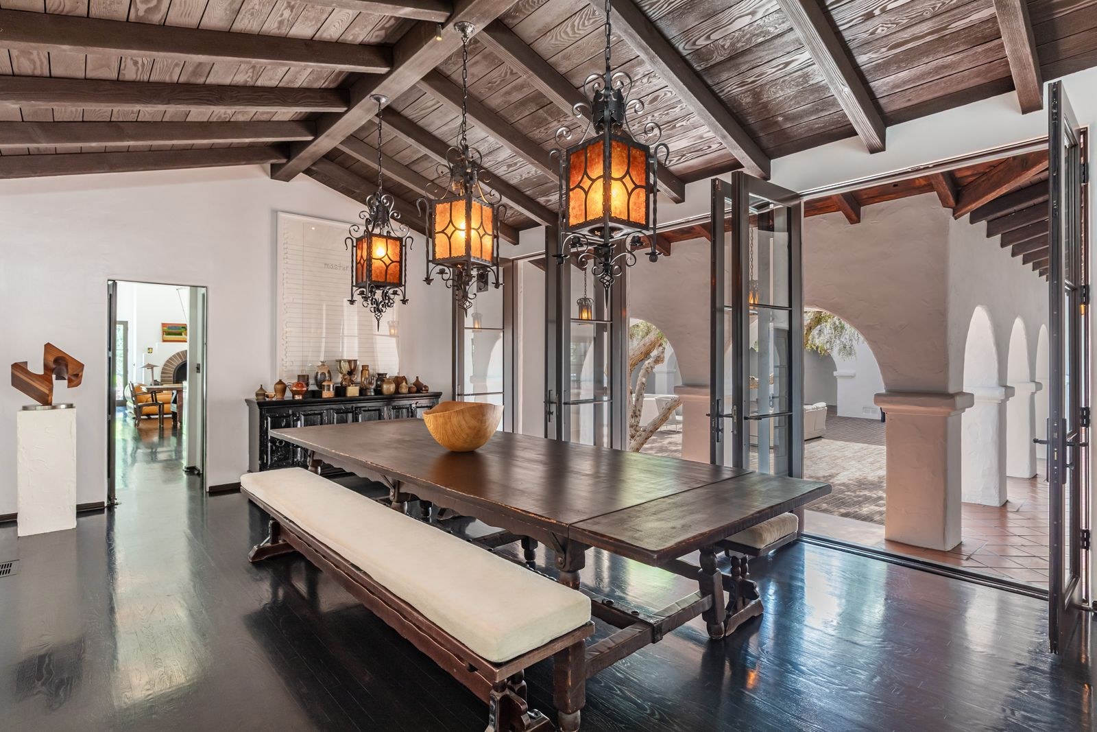At the back of the living room, a door leads you inside the formal dining room, which continues the same Spanish Colonial detailing, with dark wood floors, exposed ceiling beams, and lantern-style fixtures centered over a long table.