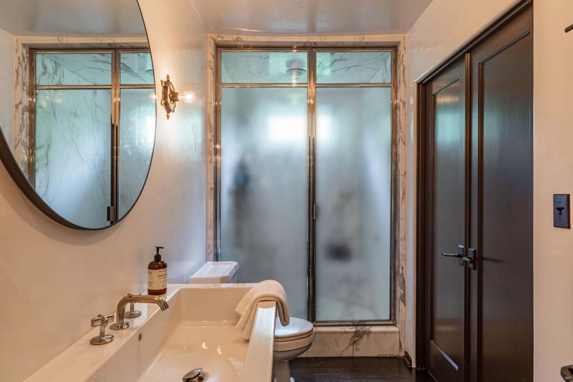 A second bathroom sits nearby with a glass-enclosed shower framed in marble, a single vanity, and more compact proportions, offering a separate, more private layout.
