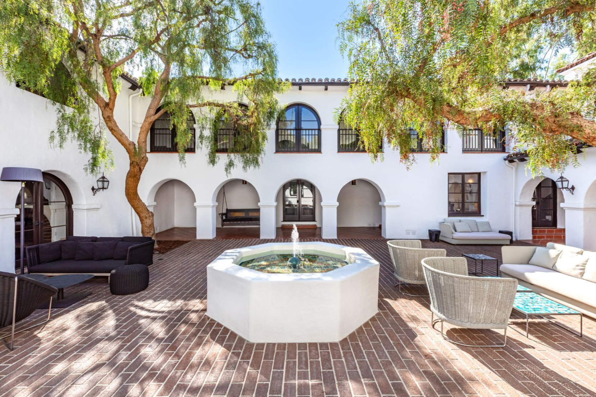 Around a fountain, multiple seating areas are arranged across the brick surface, with sofas and lounge chairs positioned beneath tall trees that provide natural shade and break up the open space.