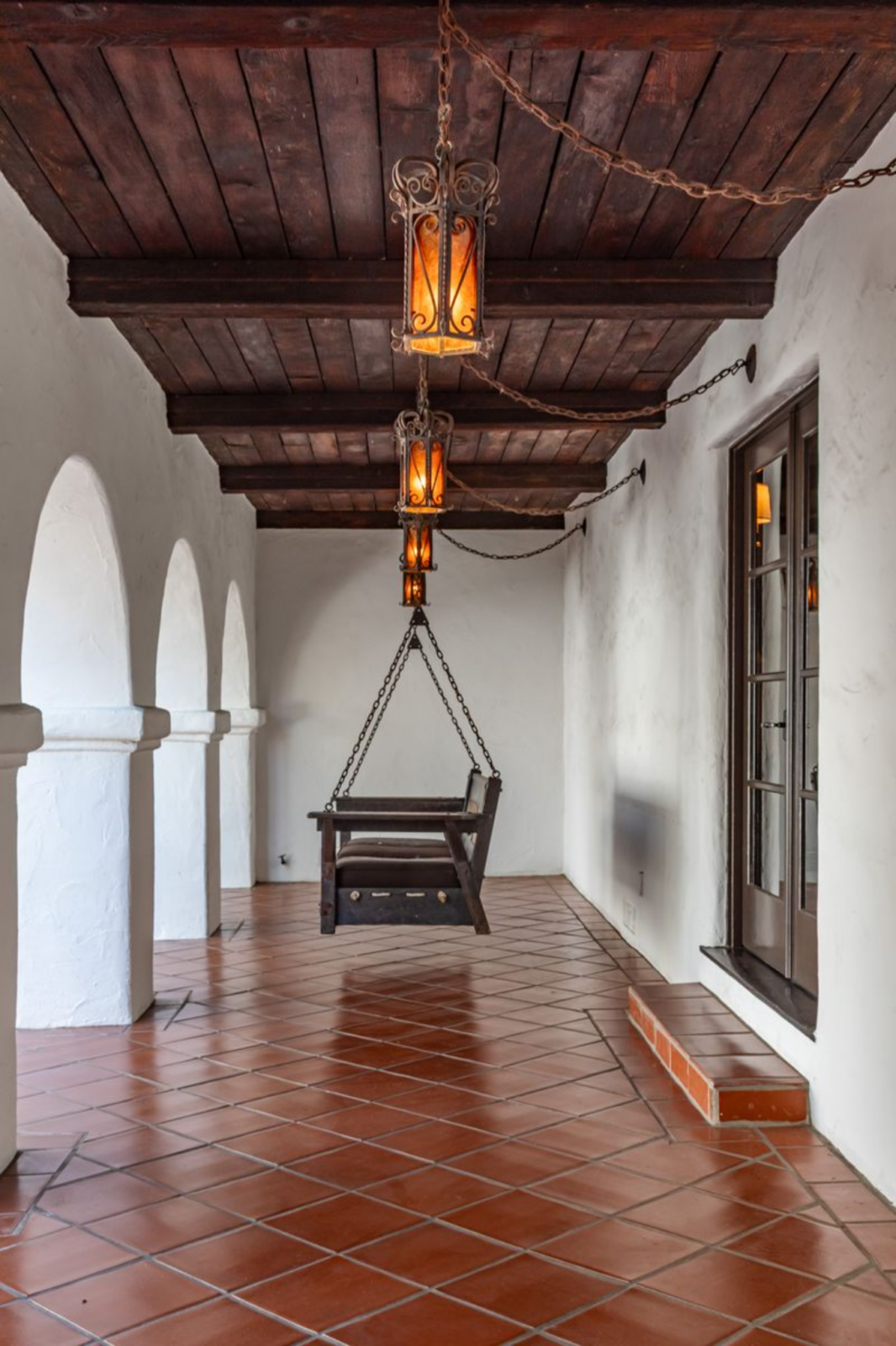 Along the perimeter, covered walkways with terracotta tile floors and wood-beamed ceilings run beneath a row of wrought-iron lanterns, creating a transition space before entering the house.