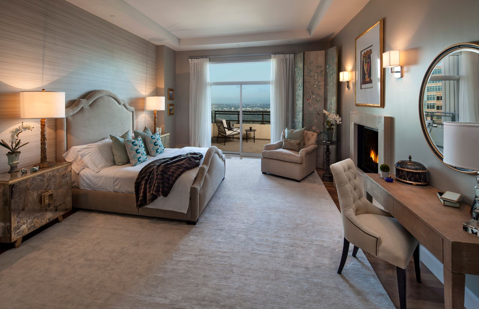 Down the hall, the primary suite has a tall upholstered bed that anchors the room against a textured wall, while layered neutrals, a fireplace, and a sitting area by the balcony doors give it a lot of functionality.