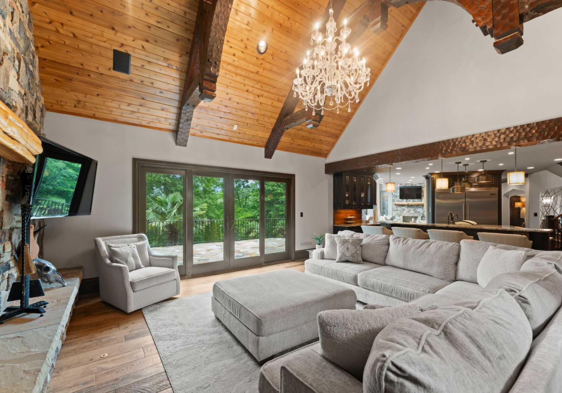 Above, the vaulted wood ceiling, exposed beams, and crystal chandelier add height and contrast, while the sectional seating and large ottoman make the room function as an everyday gathering space that connects directly to an outdoor terrace.