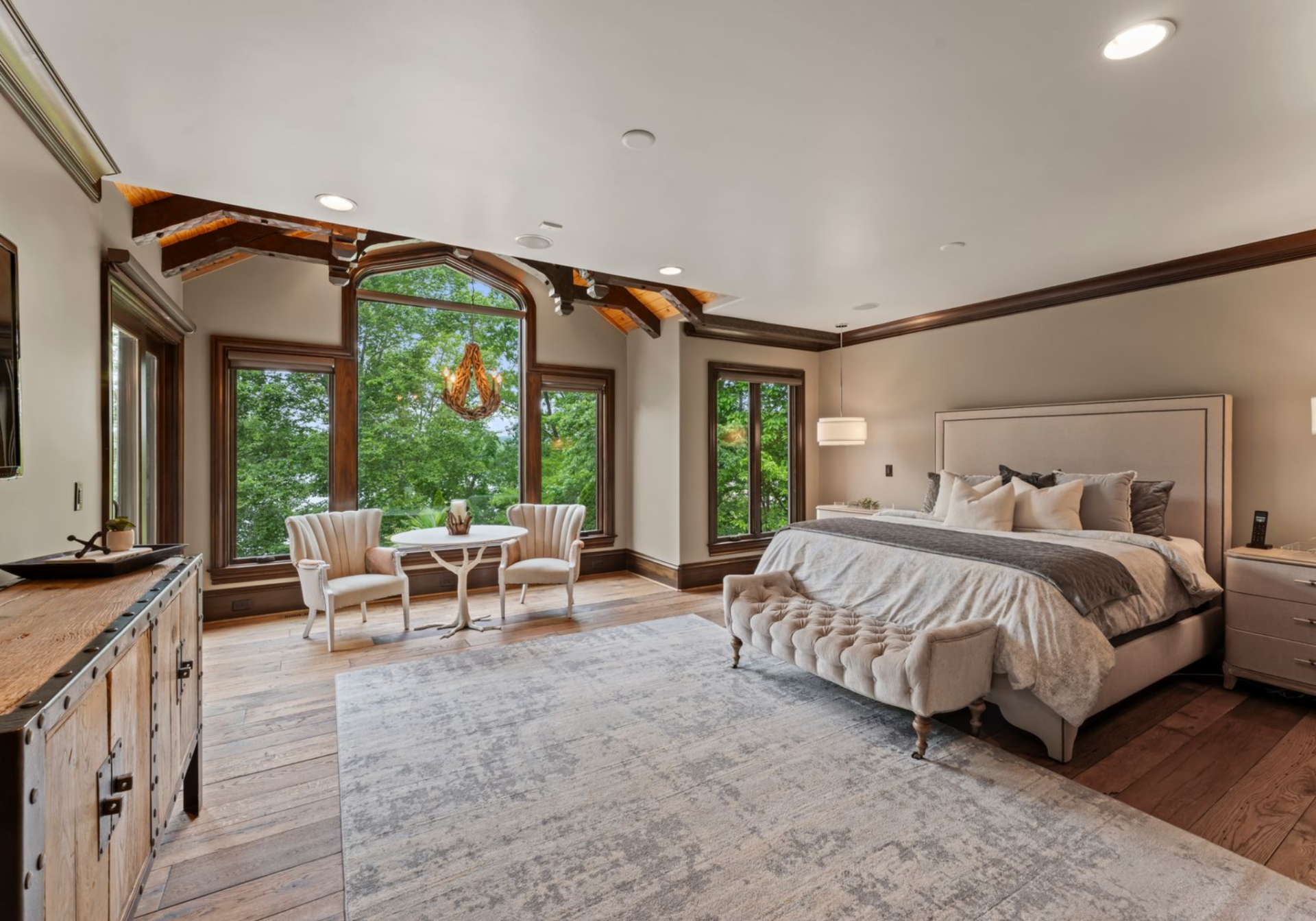 The bedroom opens with wide-plank hardwood floors and a soft, neutral palette anchored by a large upholstered bed and bench.