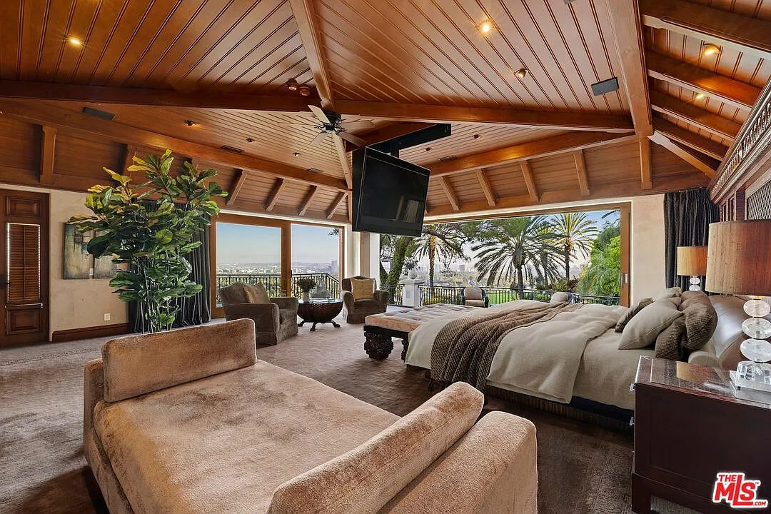 Starting with the bedroom, which features warm wood-beamed ceilings and full-wall glass doors that open to a private balcony, positioned just outside the bed and indoor lounge area.