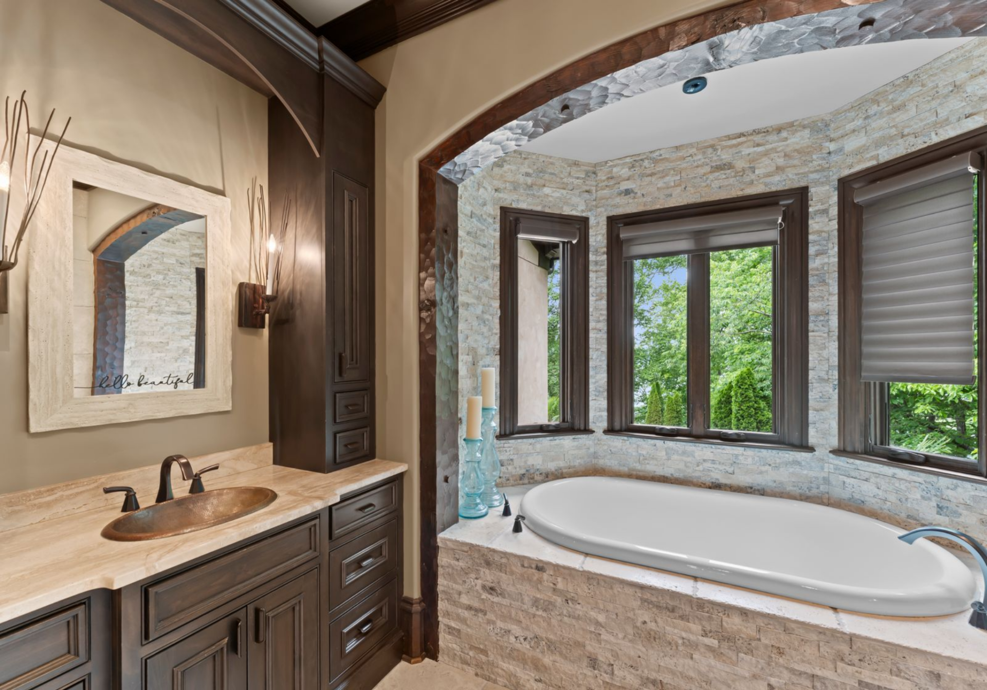 The en-suite bathroom begins with a built-in soaking tub set within a stone alcove, positioned next to one of the vanities with warm wood cabinetry, stone countertops, and a metal sink.