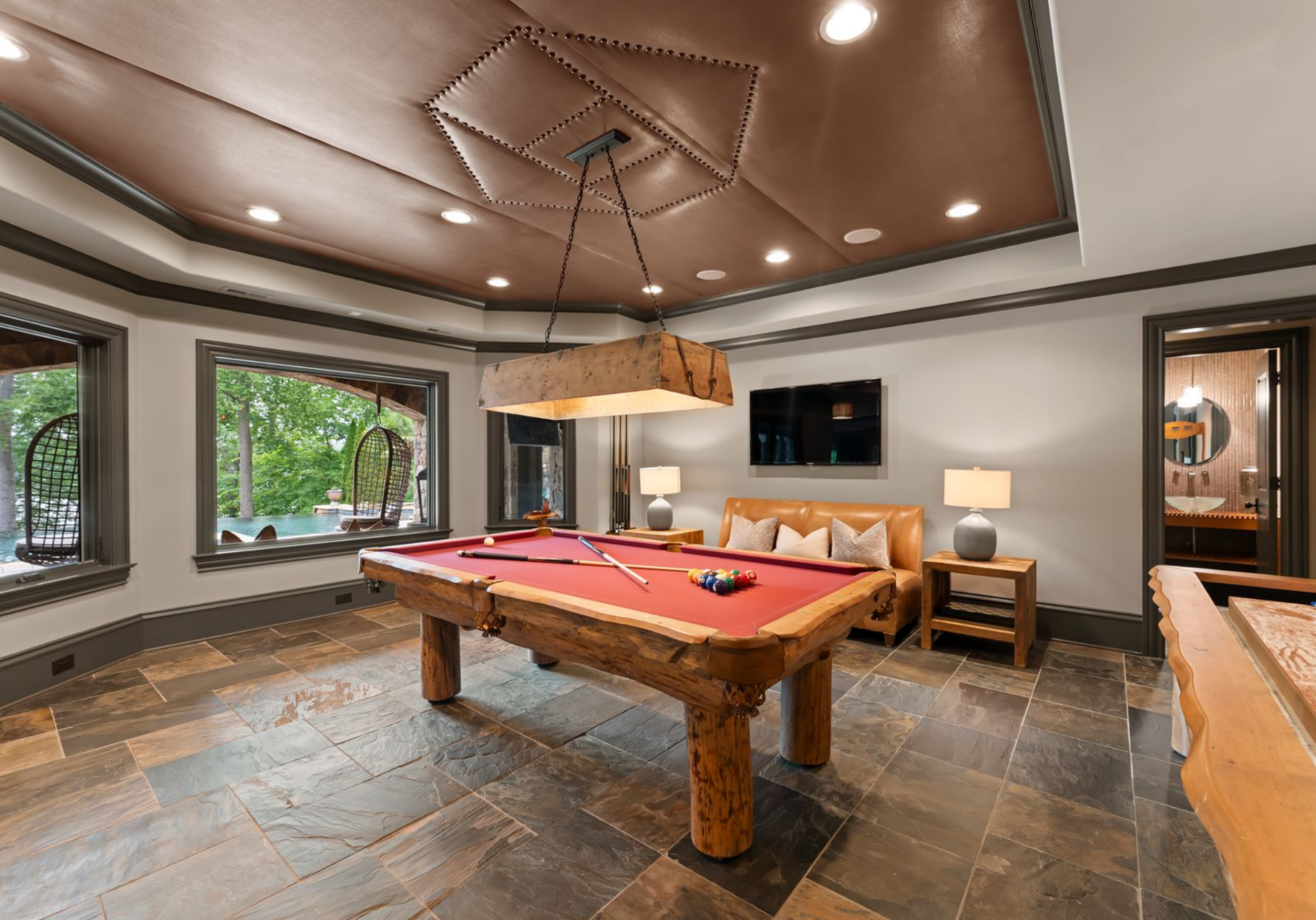 A leather-paneled ceiling, stone flooring, and a box-style hanging light give the billiards area a heavier, club-like look, while the windows keep it connected to the outdoor areas.