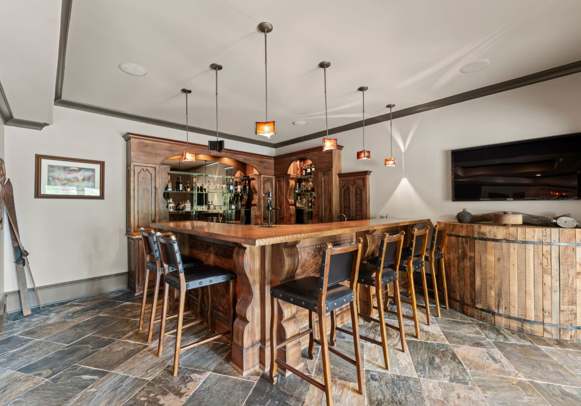 Nearby, the bar is fully built out with wood cabinetry, glass shelving, pendant lights, and seating along the counter.