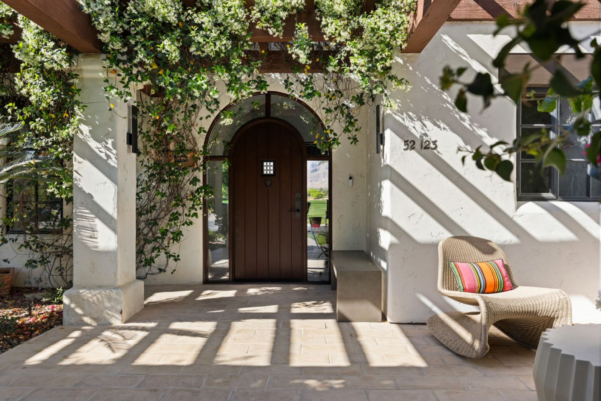 Arnold Palmer's home welcomes guests with a sun-dappled courtyard, where white stucco walls contrast with dark wood beams and lush greenery before the arched front door.