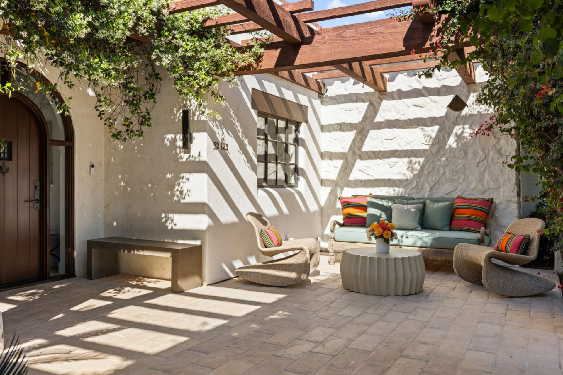 A pergola overhead casts shifting shadows on the lounge area beside the front entrance, made up of a soft blue coach and matching structural rocking chairs.