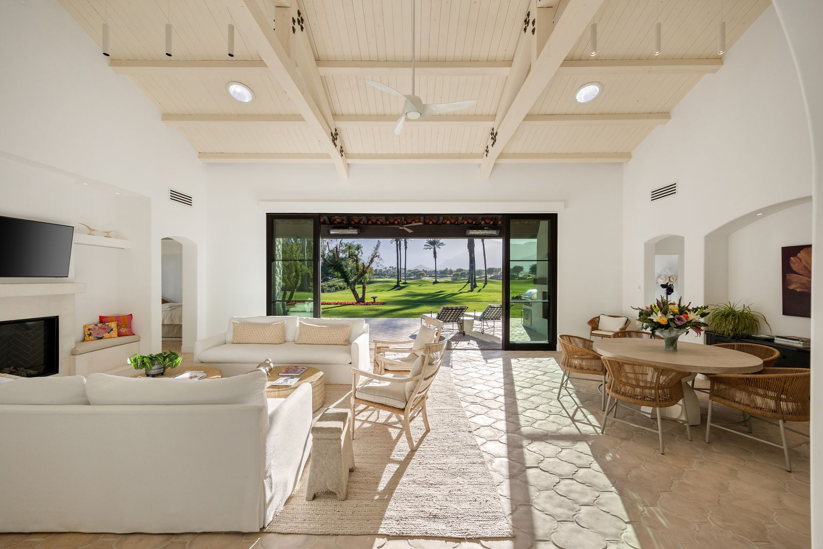 The room sits beneath a vaulted, white-painted beam ceiling, creating an open and inviting design, complemented by a neutral palette of soft furnishings, woven textures, and warm-toned stone flooring.