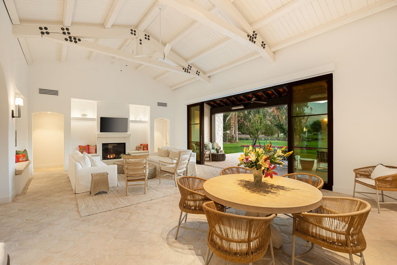 On the other side of the room, the dining area is tucked into an alcove, accented by soft lighting and a colorful centerpiece of flowers on top of the rounded light wood table.