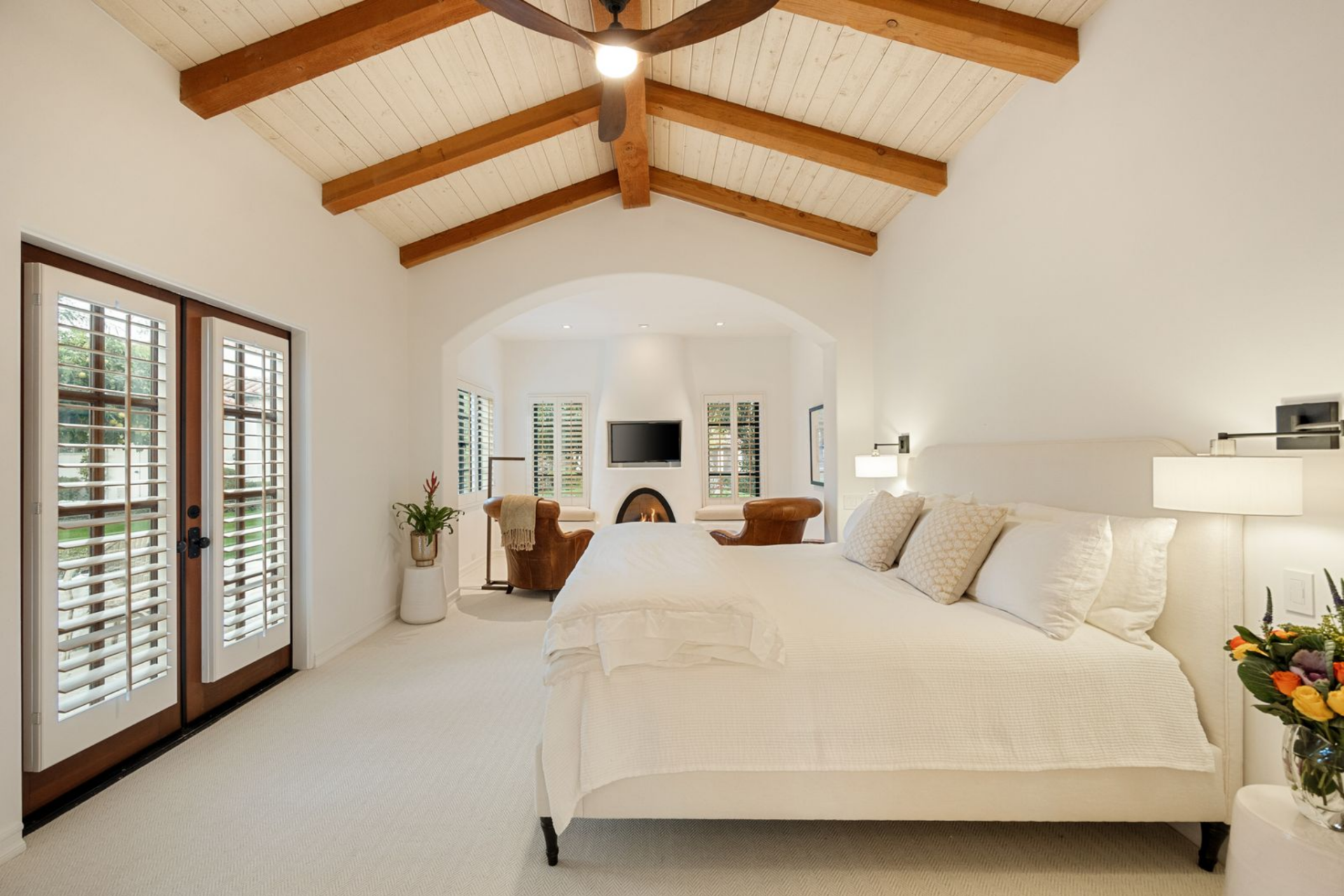 The bedroom opens beneath a vaulted, wood-beamed ceiling, giving the room height without overwhelming the simple white palette.