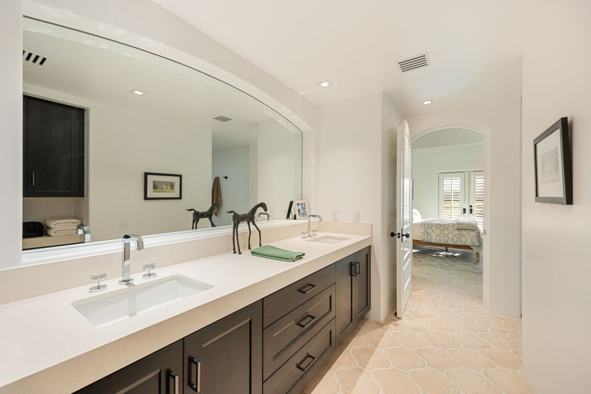From the bedroom, the suite connects to a long, light-filled bathroom with patterned tile floors and a double vanity set beneath an oversized arched mirror.