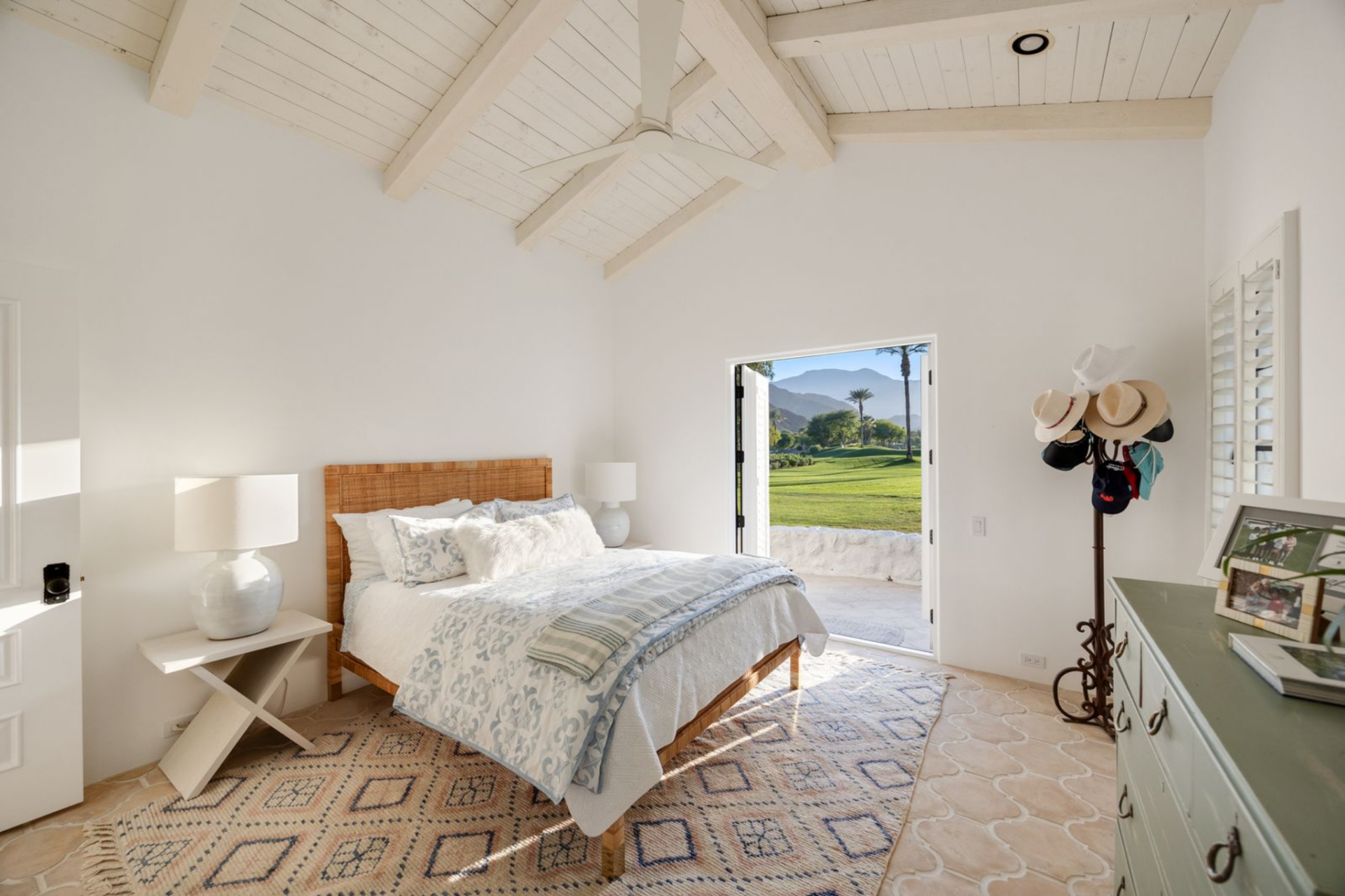 The room is simple, with a woven bed frame, a patterned rug, and an open doorway that looks out toward the golf course and mountains.