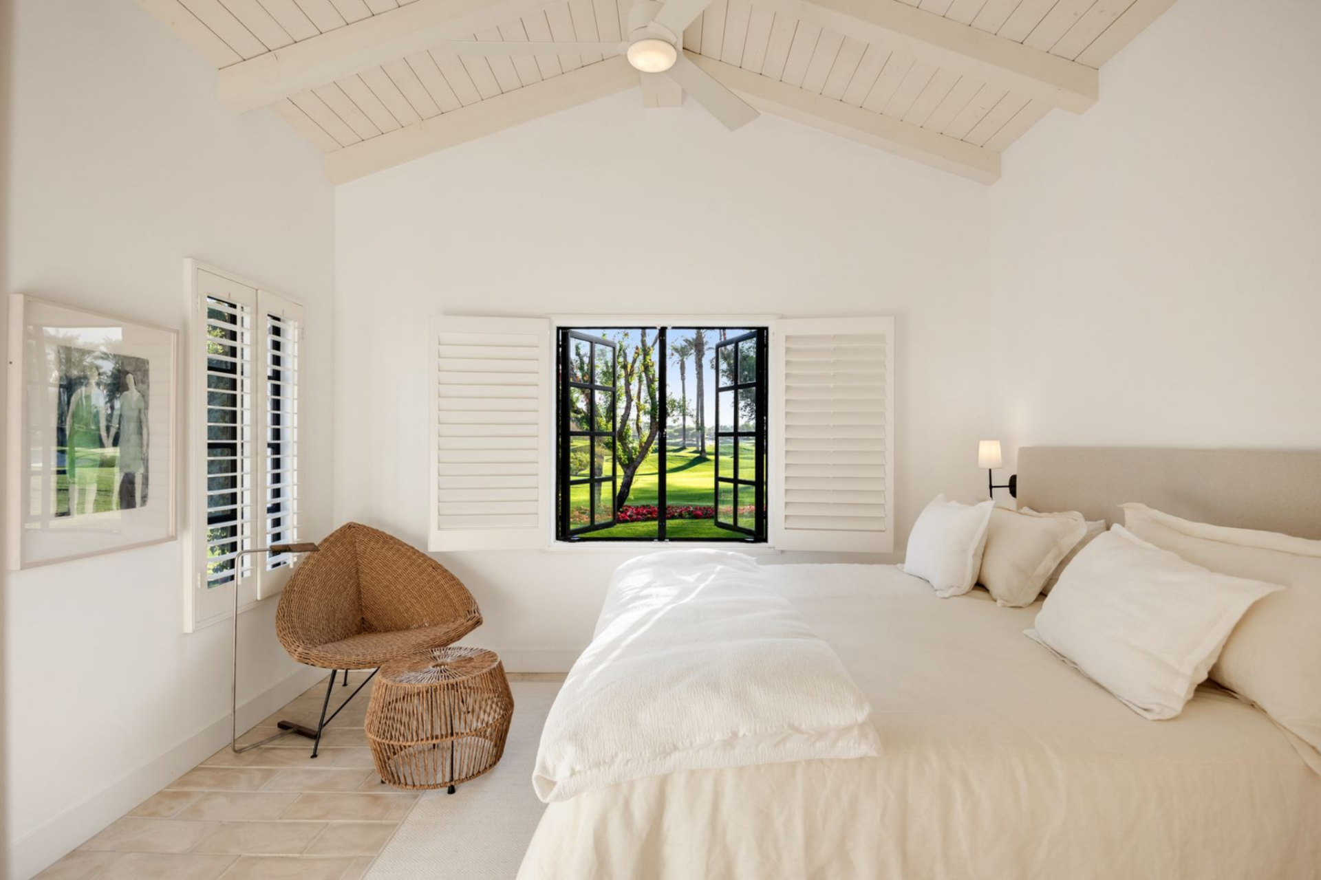 Another bedroom continues the all-white palette, with a high ceiling, shuttered windows, and a small sitting area near the corner.