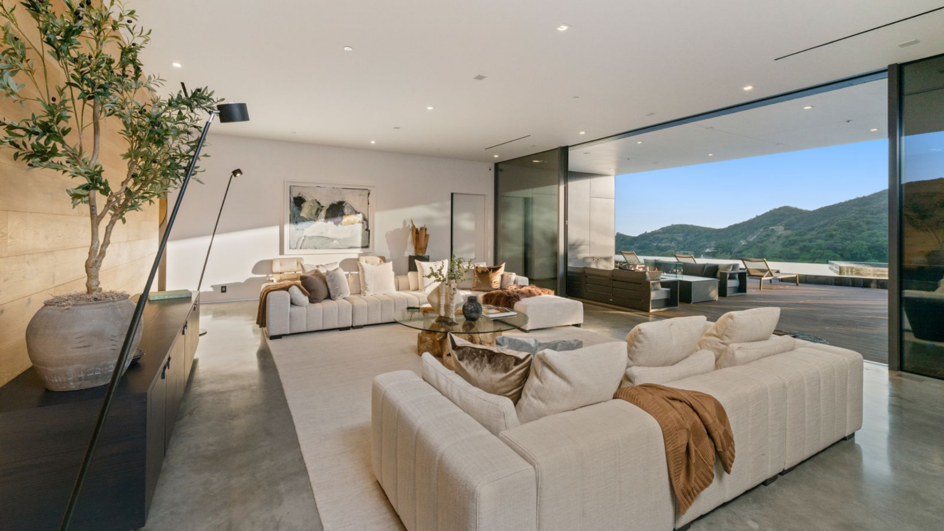 The layout consists of a series of low-profile sectional sofas arranged around a glass coffee table with a sculptural wood base, creating a central gathering area without interrupting the views.