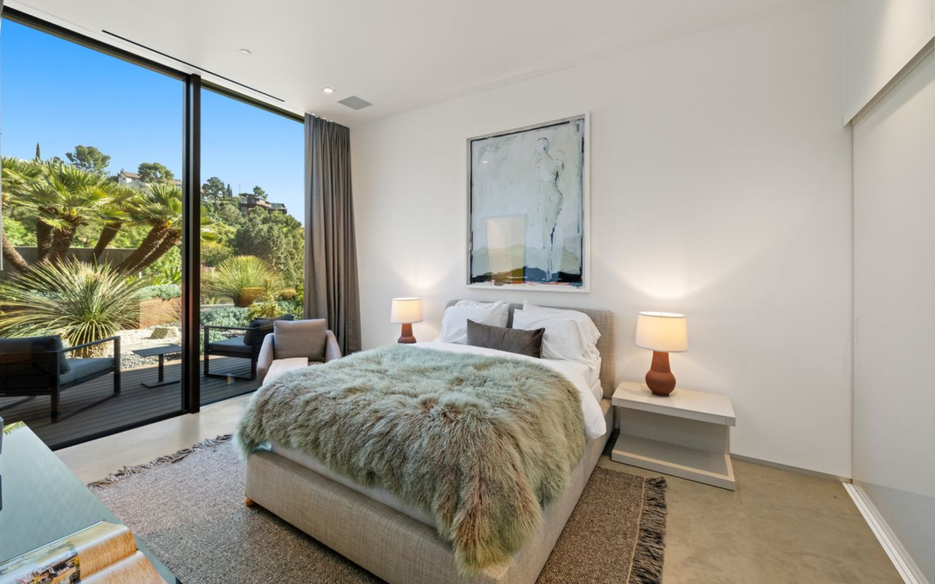The room uses a softer palette, with neutral bedding, a textured throw, and a compact seating area near the glass.