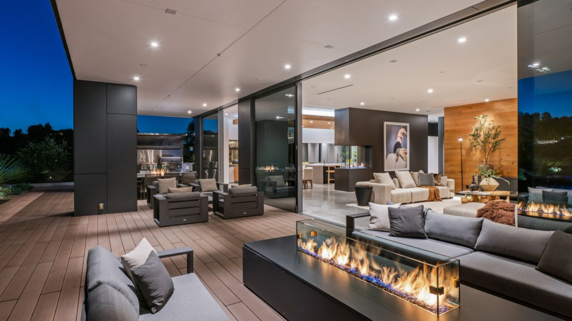 Sliding glass walls connect the living room and kitchen to multiple outdoor seating zones, including a fire pit positioned just outside the glass.