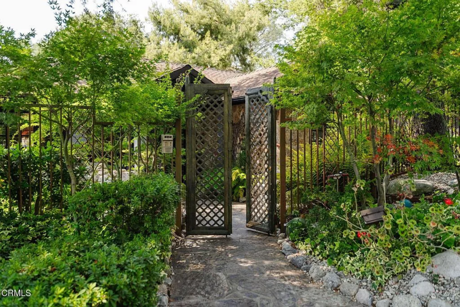 A narrow stone path moves through layered greenery and low rock borders before arriving at a more defined front courtyard, where the house just slightly comes into view.