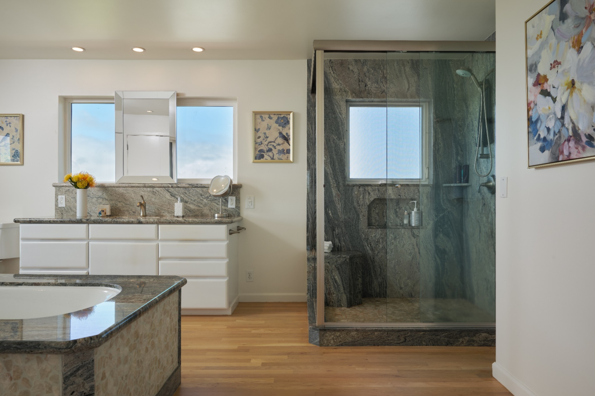Inside the bathroom, a white vanity with stone counters sits beside a glass-enclosed shower wrapped in dark stone, while a built-in soaking tub sits in the center of the room, lined in matching stone.