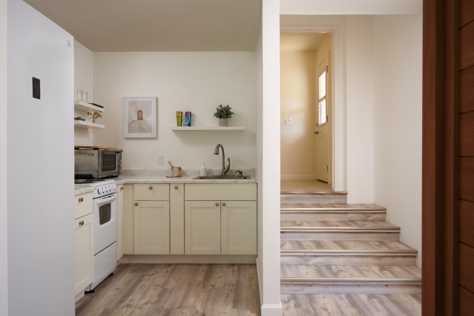 Just inside, the suite includes a compact full kitchen with cream cabinetry, a white range, open shelving, and a small sink set beneath simple wall-mounted storage. 