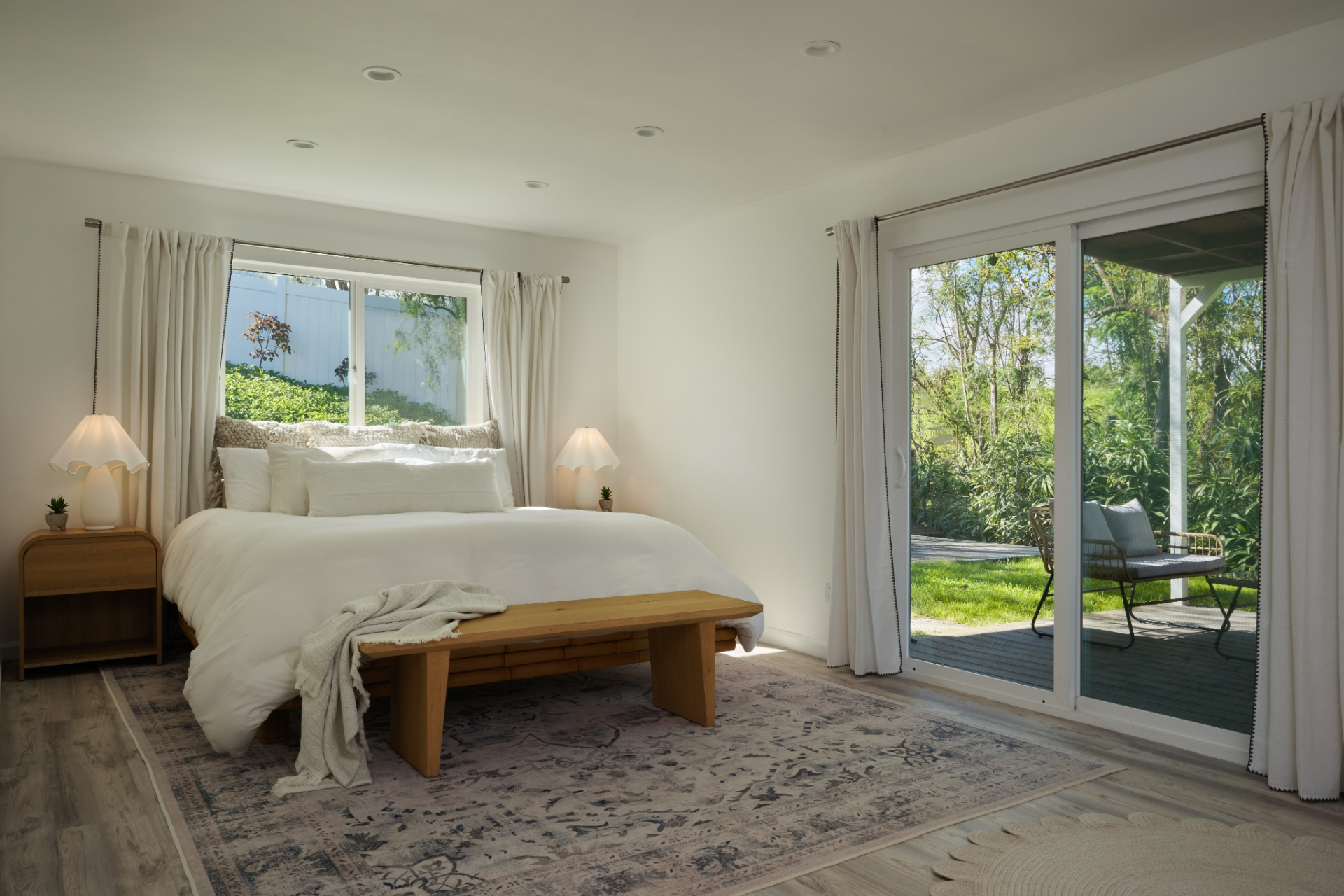 The bedroom is bright and simply designed, with light flooring, white bedding, and sliding glass doors that open directly to a private yard.