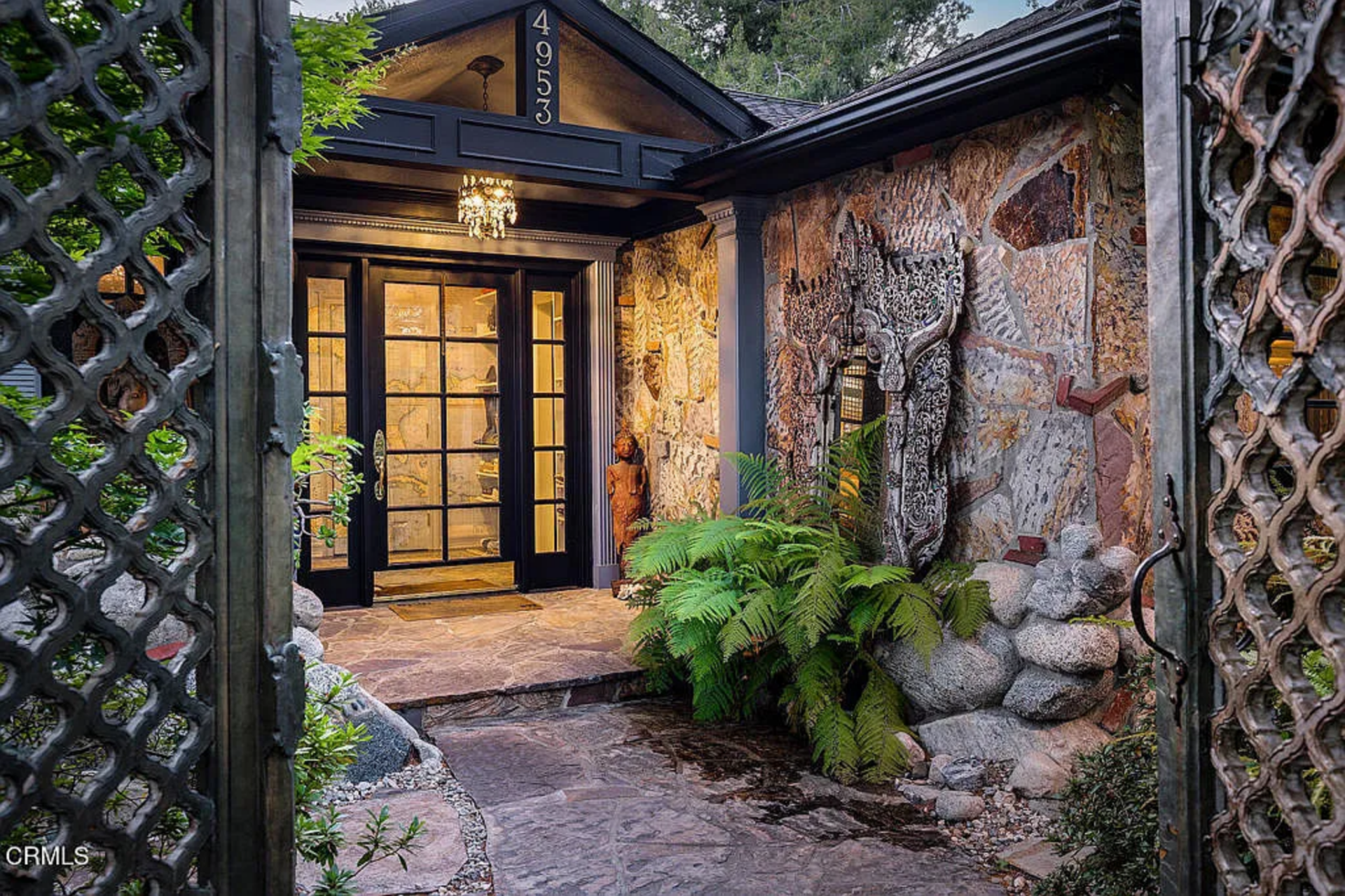 At the end of the path, dark-framed glass doors sit beneath a peaked porch with a hanging light, while the surrounding exterior mixes stone, wood trim, and carved decorative elements.