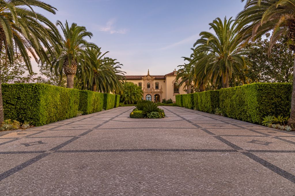 The entrance to the main house begins with a long, patterned motor court framed by tightly clipped hedges and palm trees, guiding you directly toward a symmetrical façade finished in warm stucco and stone.