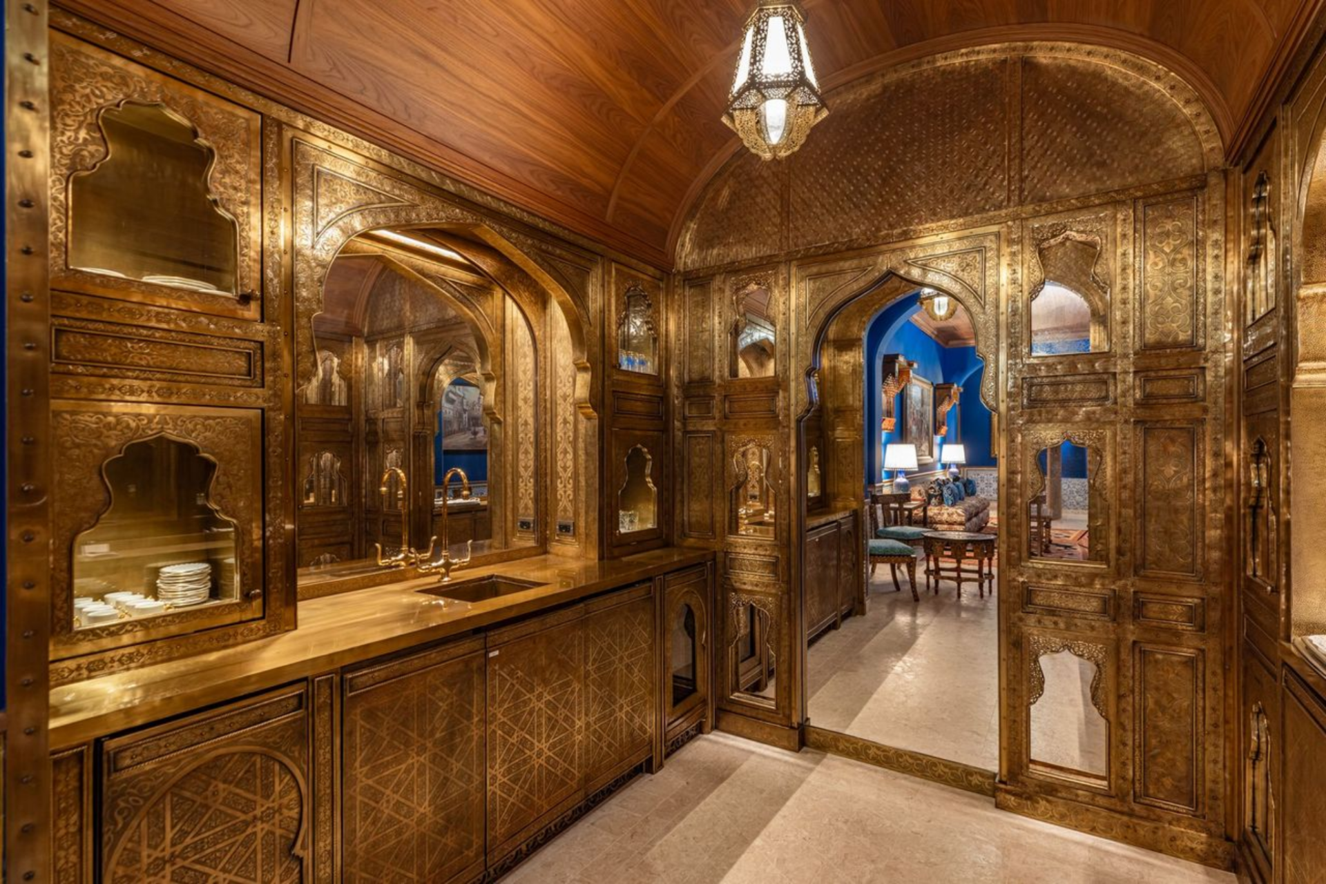 Engraved brass cabinetry, arched doorways, mirrored panels, and warm lantern lighting create a jewel-box transition from the blue sitting room into the bar area.