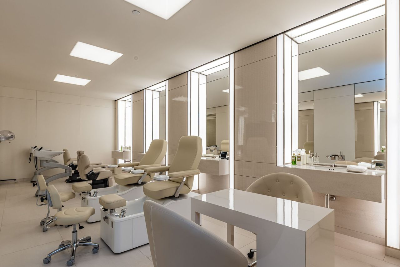 The beauty salon is bright with backlit mirrors, white counters, and multiple styling and treatment stations for manisures, pedicures, and hair styling.