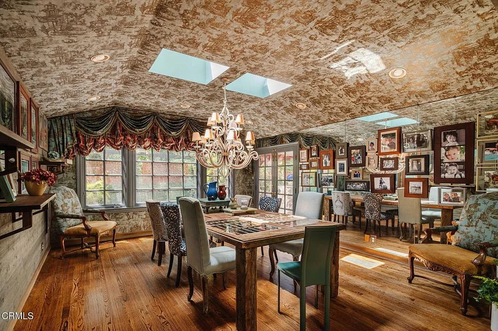 The walls and sloped ceiling are wrapped in the same printed wallpaper, broken up by skylights overhead, while a chandelier drops over a long rustic table surrounded by a mix of upholstered dining chairs rather than a matching set.