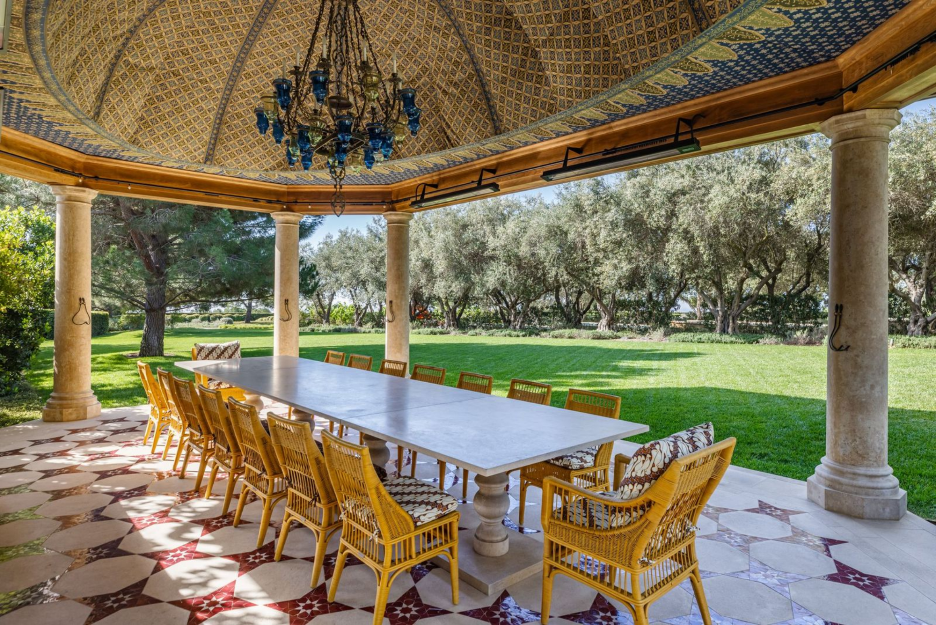 Within the space, a long central table is surrounded by woven chairs, while overhead heaters and a chandelier make the space usable across seasons.