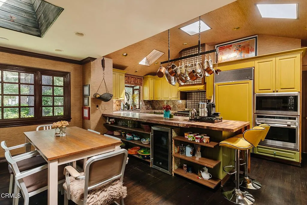 In the next room, the kitchen features a long prep island that sits at the center with a butcher-block top and two yellow swivel stools, while a second wood table extends off to the side for casual meals.