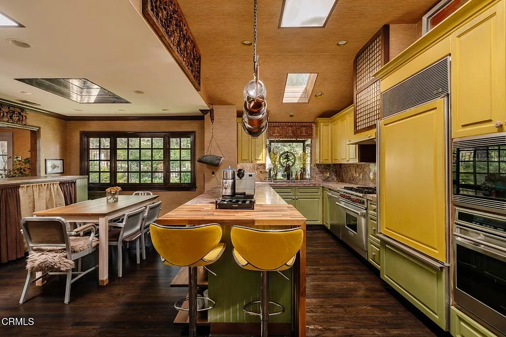 Overhead skylights brighten the ceiling, a hanging pot rack drops above the island, and the range, sink, and built-in ovens are spaced along the walls.