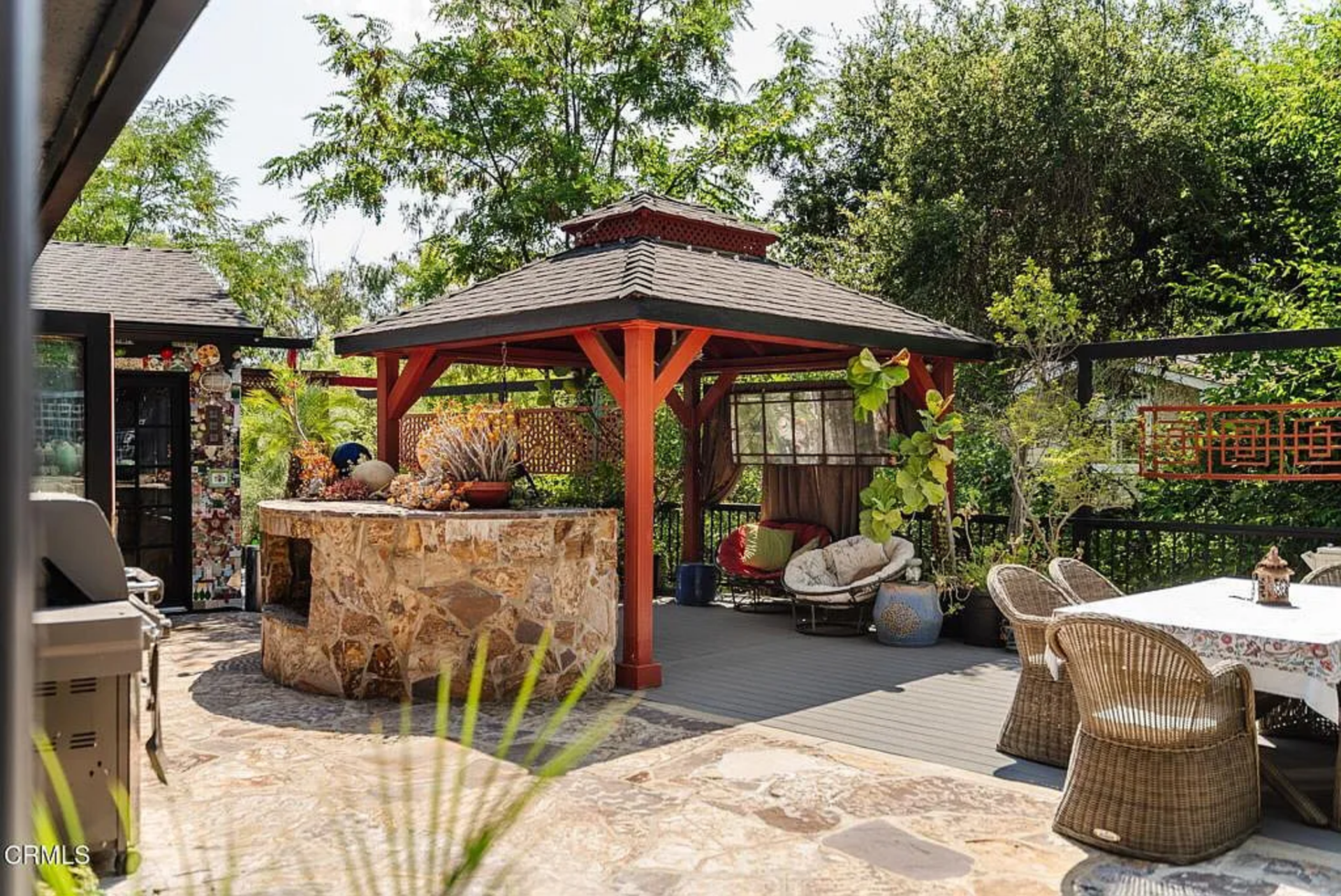A red wood pavilion creates one section, with a built-in stone bar set just beside it, while the covered area holds a dining table and lounge seating beneath the pitched roof.