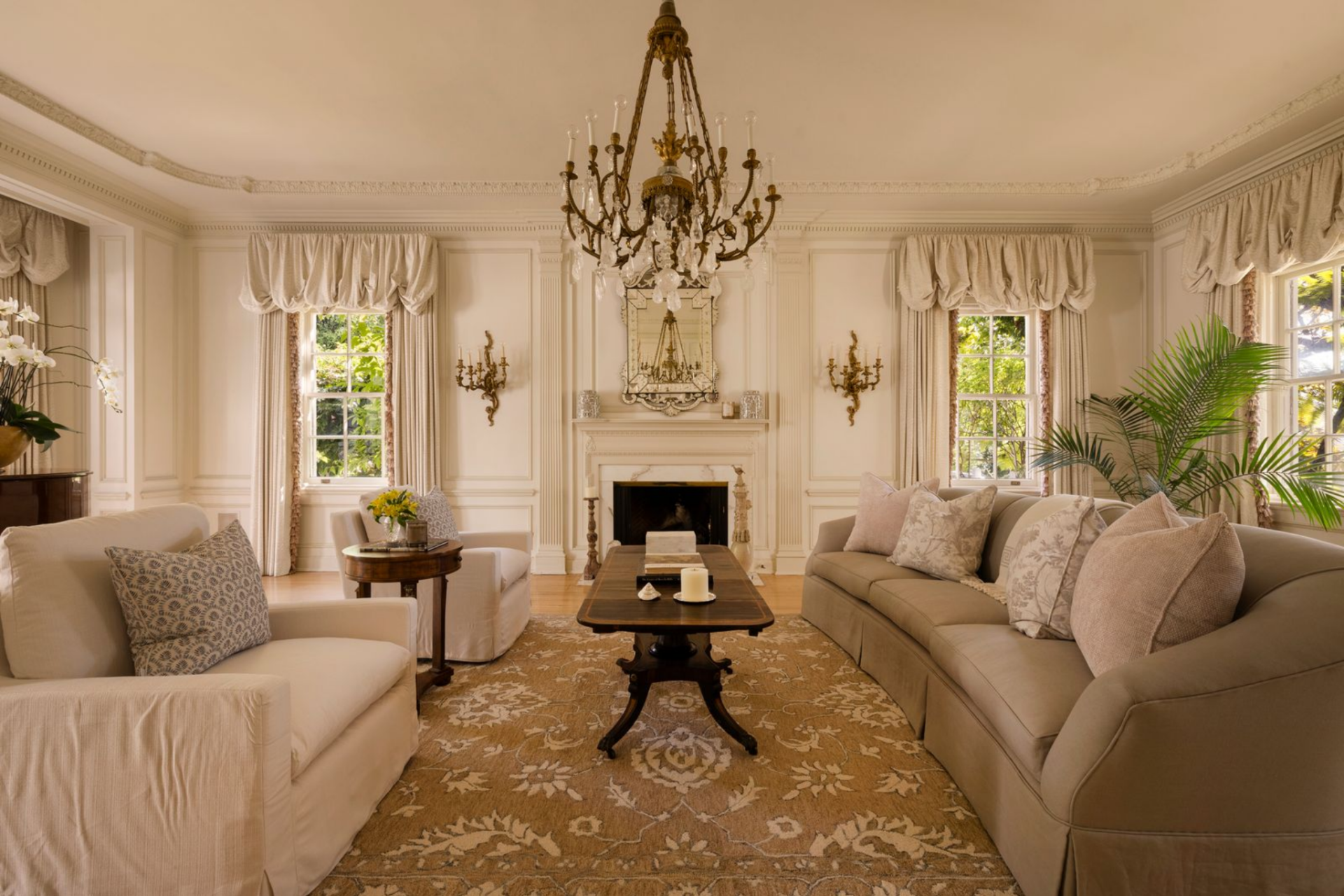 The layout is arranged in a traditional, balanced way, with the fireplace at the center, matching sconces on either side, and seating set around a long wood coffee table over a large patterned rug. 