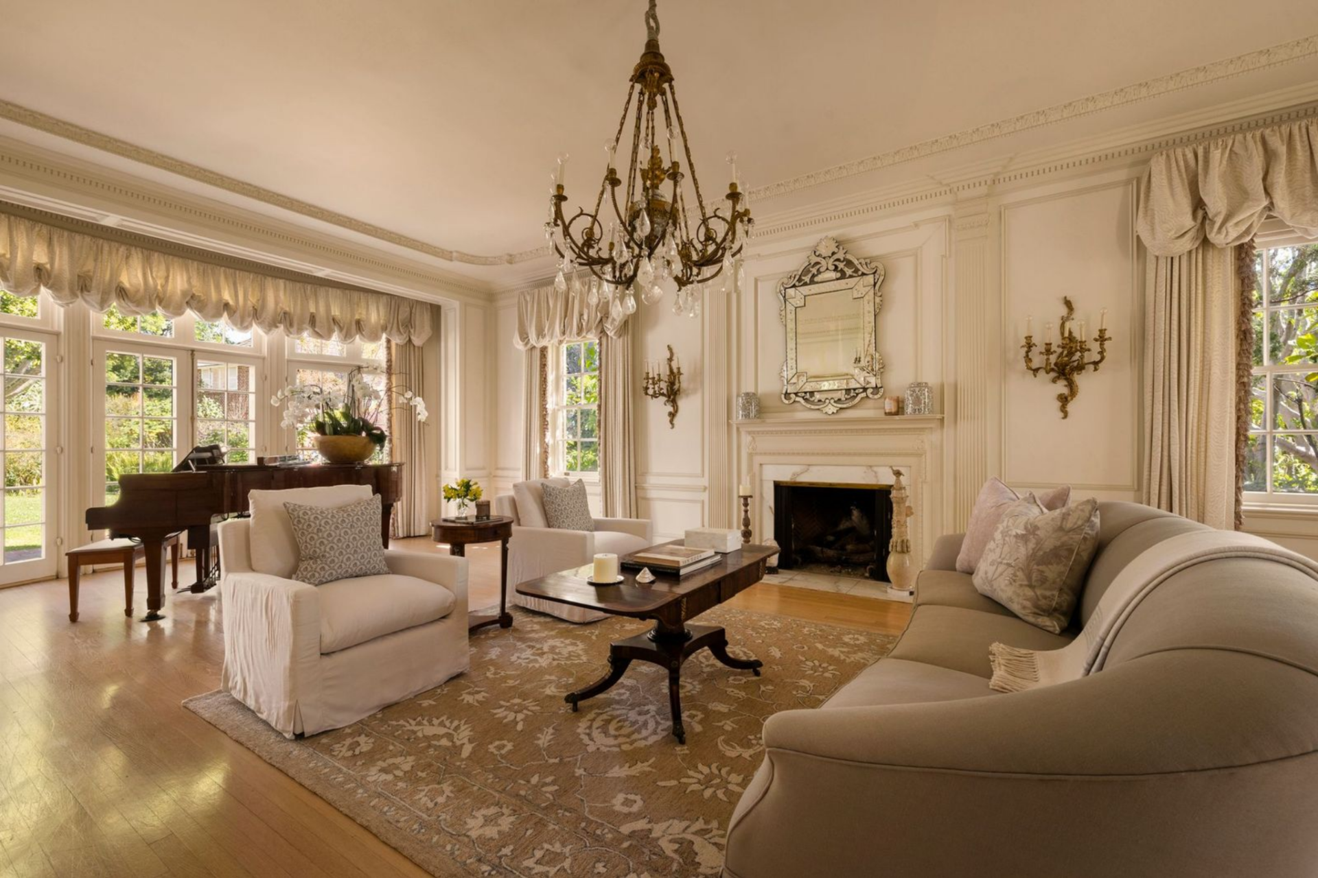 Tall windows and French doors bring in garden views along one side, and a piano sits at the far end, reinforcing the room’s formal use while keeping the flow open into the next part of the house.