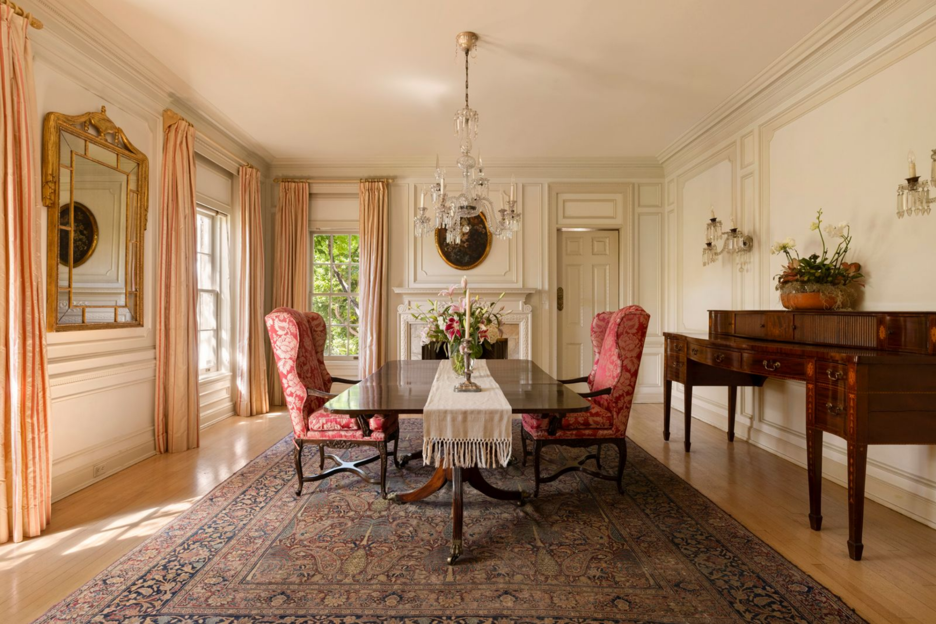 A crystal chandelier hangs over the center table, while matching sconces, an antique mirror, and a finely detailed sideboard reinforce the room’s traditional layout.