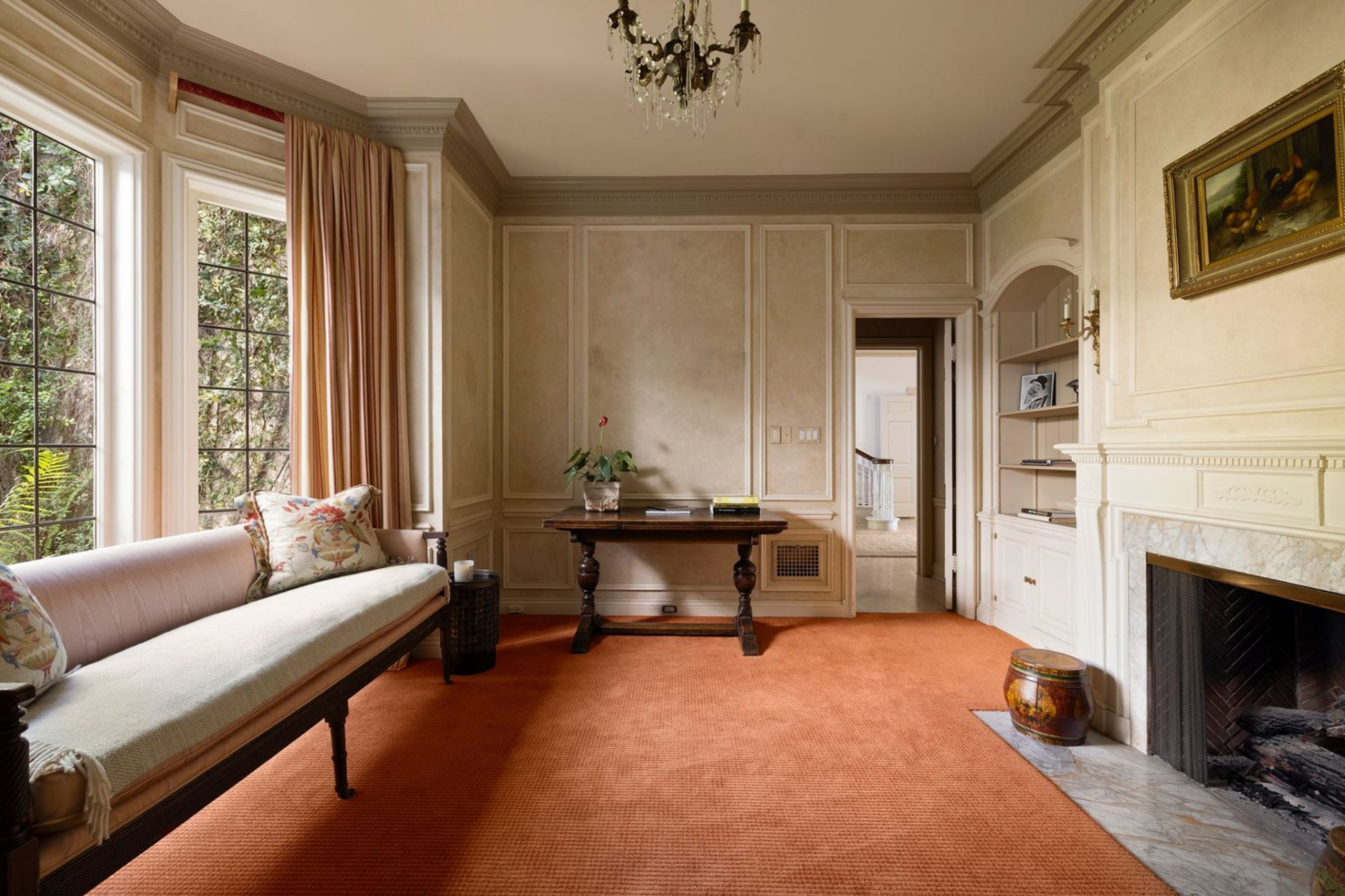 Tall windows bring light across the soft wall panels and orange carpet, while the marble fireplace, carved trim, and delicate chandelier keep the room rooted in the house’s 1930s detailing.