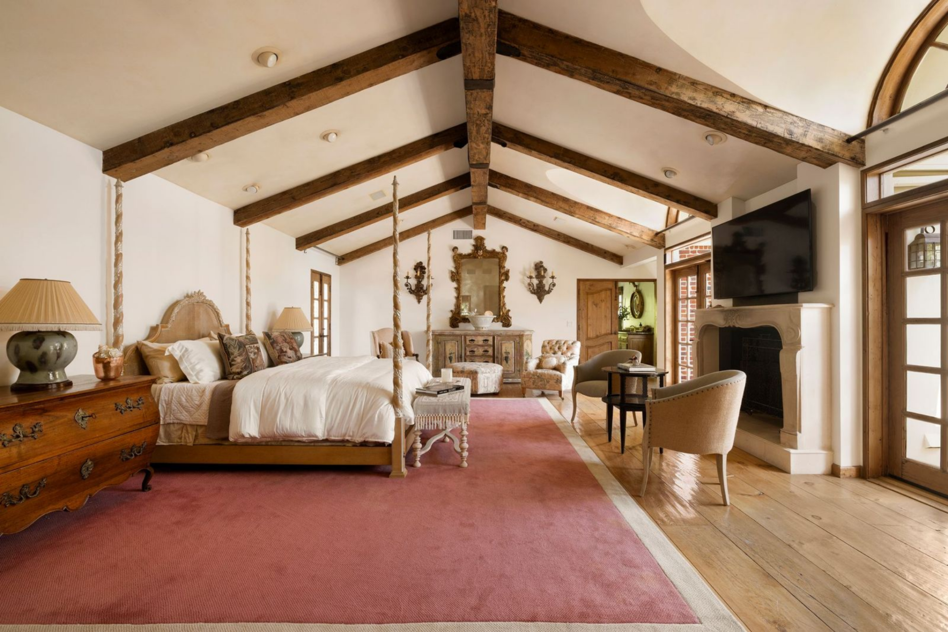 A four-poster bed sits over a large area rug across from a large fireplace with multiple doors and windows spread outward around it.