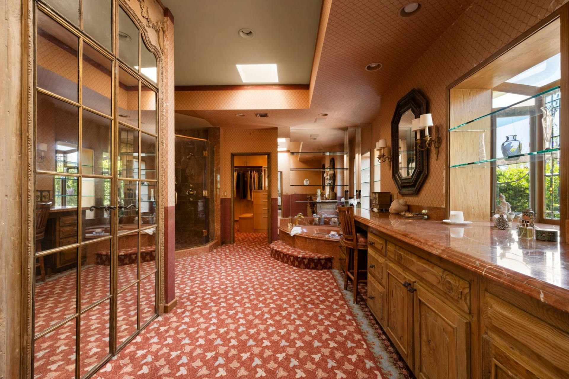 One is arranged like a dressing room and bath combined, with long wood vanities, mirrored cabinetry, patterned wallcovering, and a built-in soaking tub set into a raised platform before the shower and closet. 