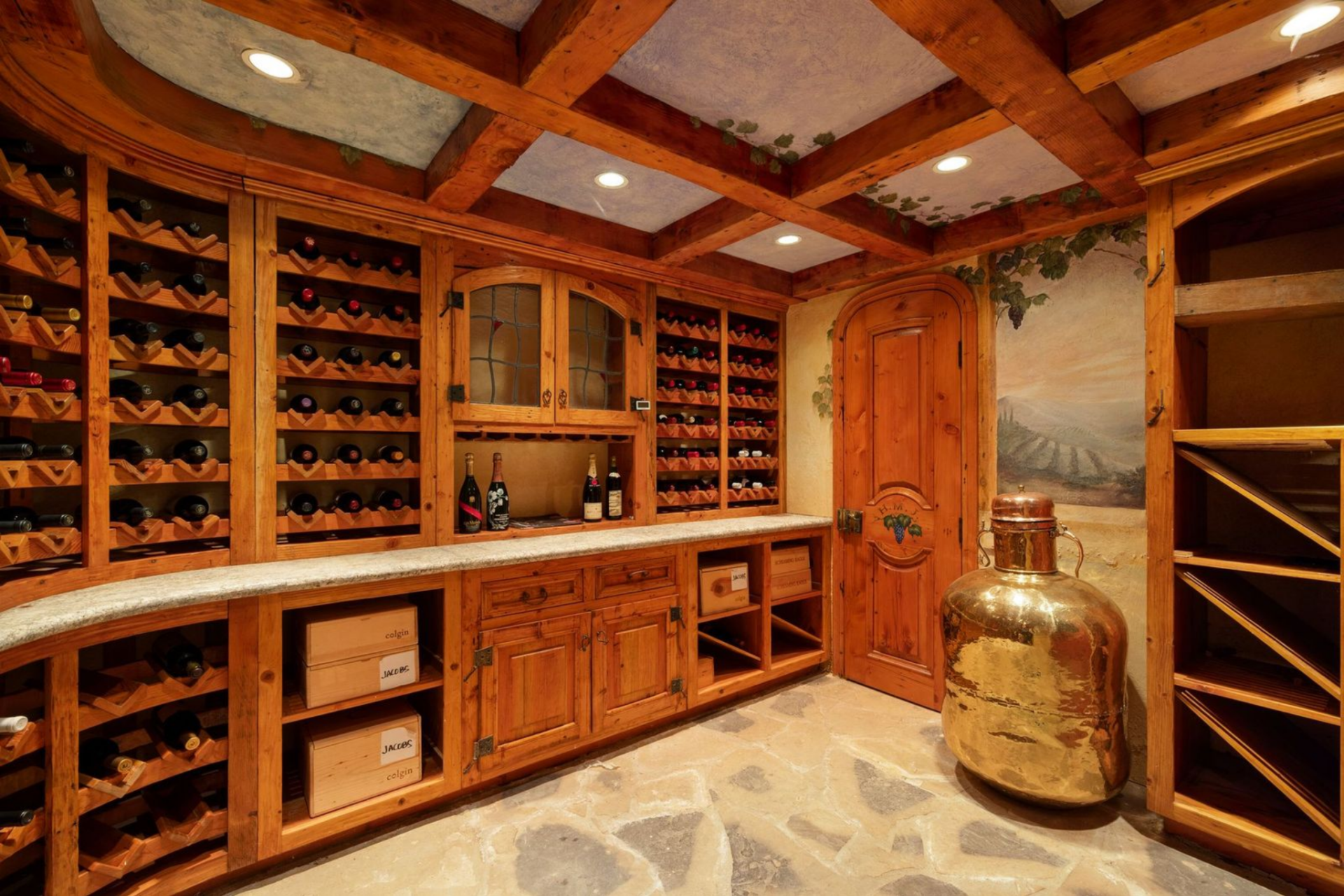The wine cellar is finished almost entirely in warm-toned wood, with bottle storage built into nearly every wall and additional cabinetry running below a stone countertop. 
