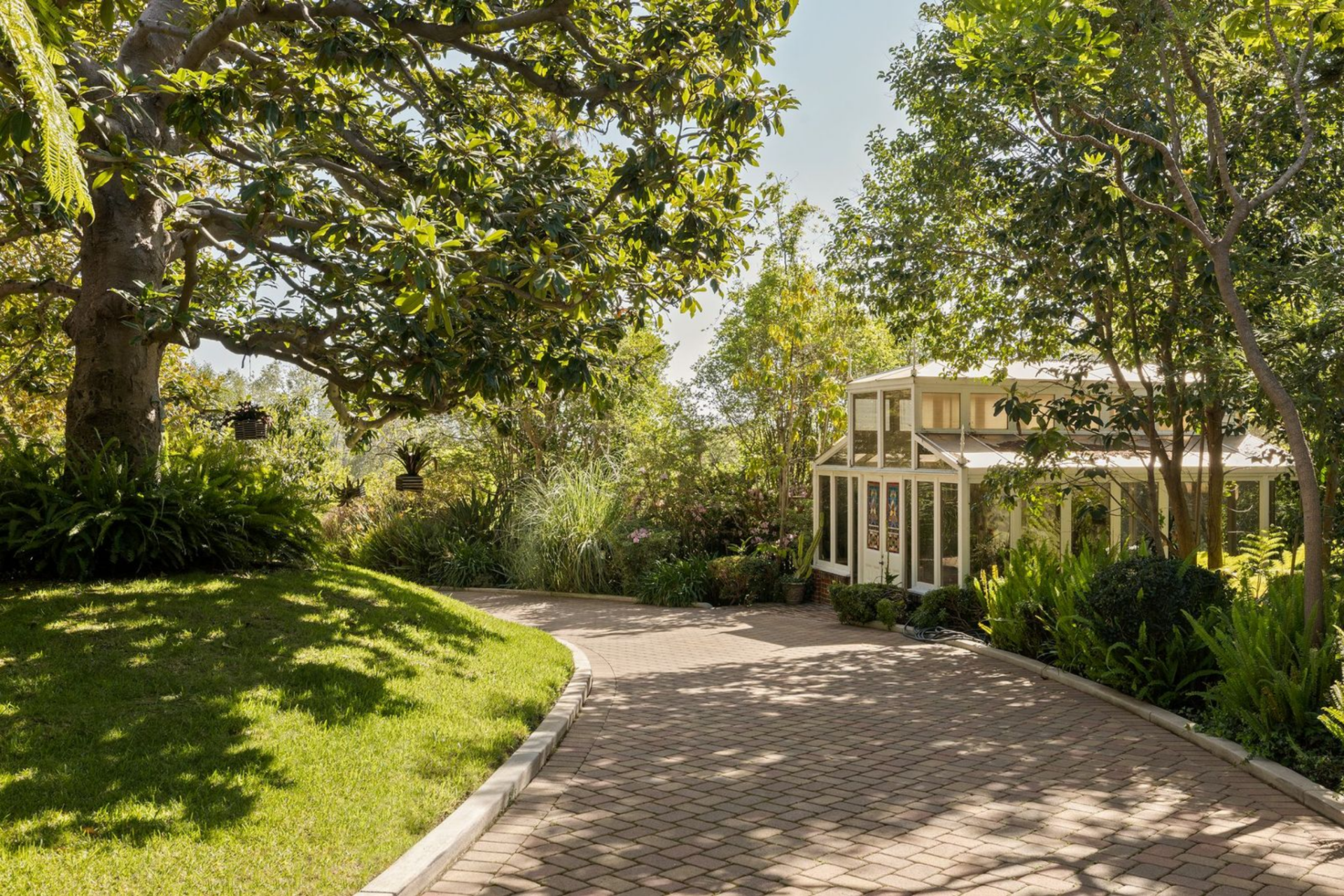 Elsewhere on the property, a winding paved path moves through dense landscaping toward one of the two greenhouses, which is tucked into the trees and finished with tall glass walls and a peaked roofline. 