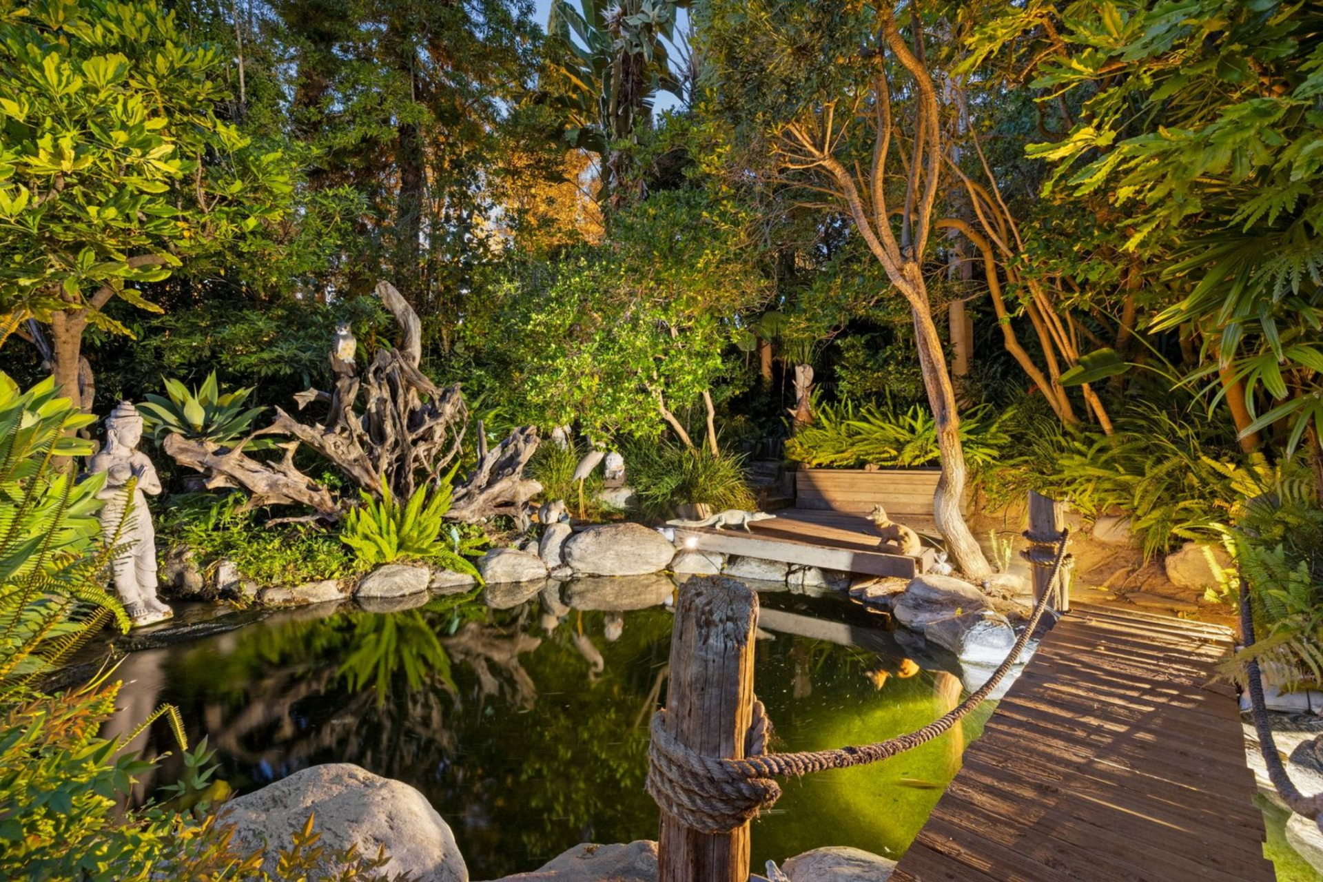 A narrow wood bridge crosses one side, and the pond is arranged almost like a small hidden garden, tucked beneath taller trees and enclosed by foliage on nearly every edge.