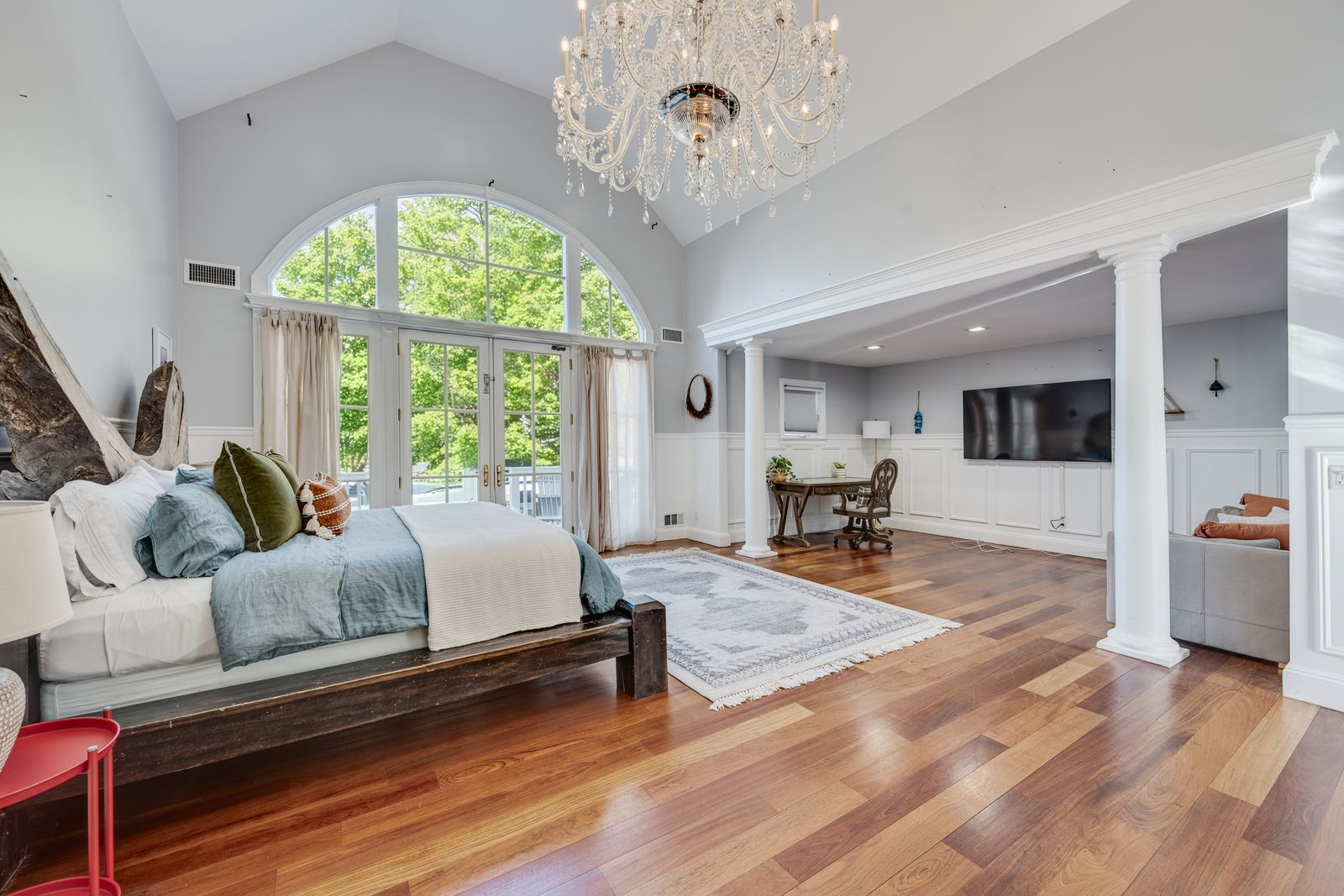 An oversized arched window and French doors bring in natural light at one end, while the sleeping area opens into a private sitting area framed by columns.