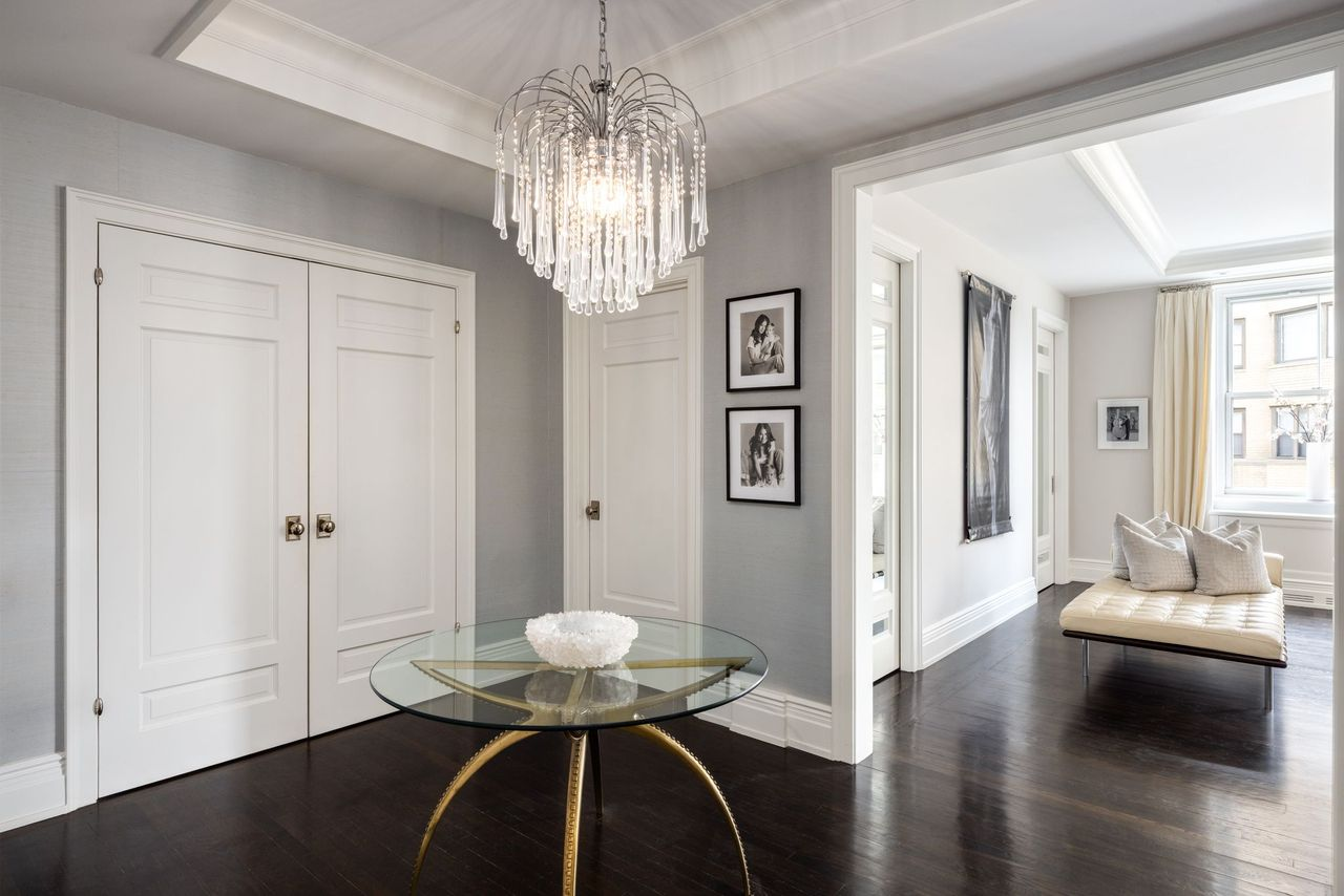 Dark hardwood floors run beneath soft gray wall coverings and crisp white molding, while a cascading crystal chandelier hangs above a round glass table positioned at the center of the space.