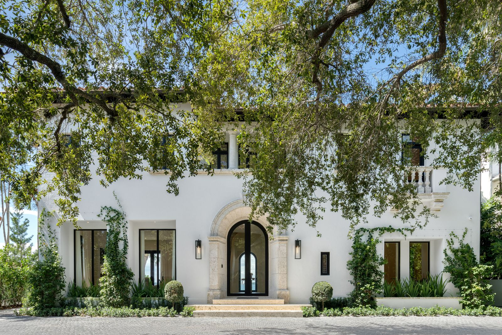 The white stucco exterior is softened by climbing greenery and symmetrical plants, while the second-floor balconies and column details subtly reference classic European architecture.
