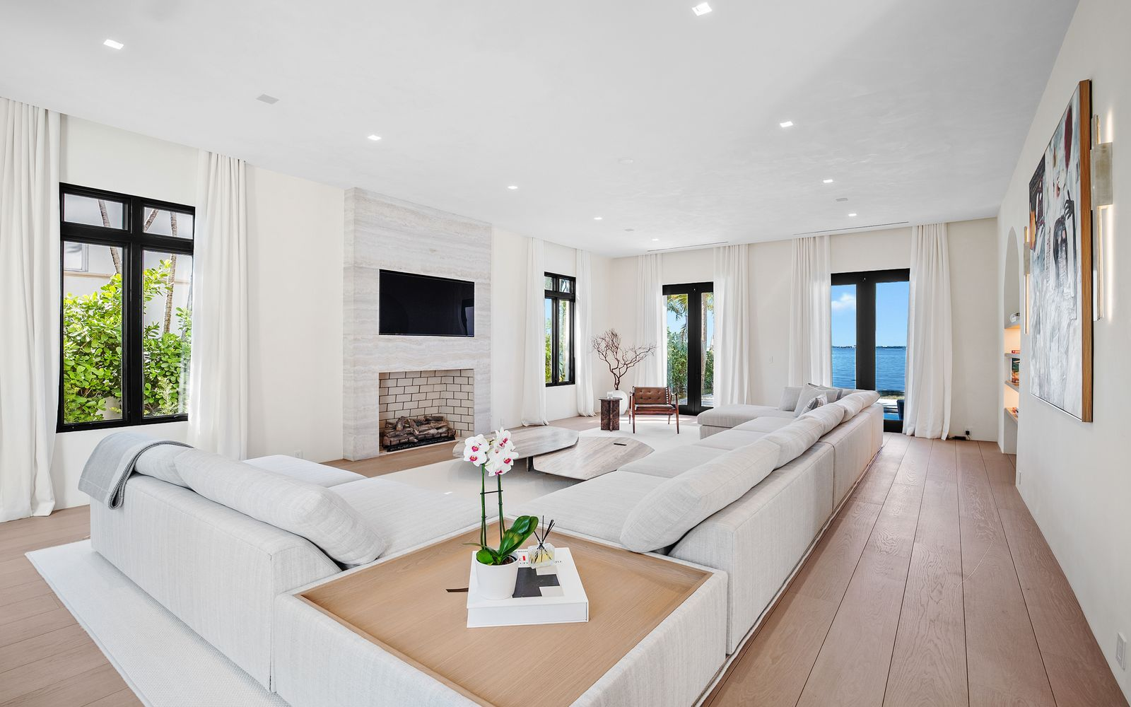 Stepping past the curved staircase, the living room opens into a long waterfront-facing space defined by high ceilings and oversized seating arranged around a travertine fireplace.
