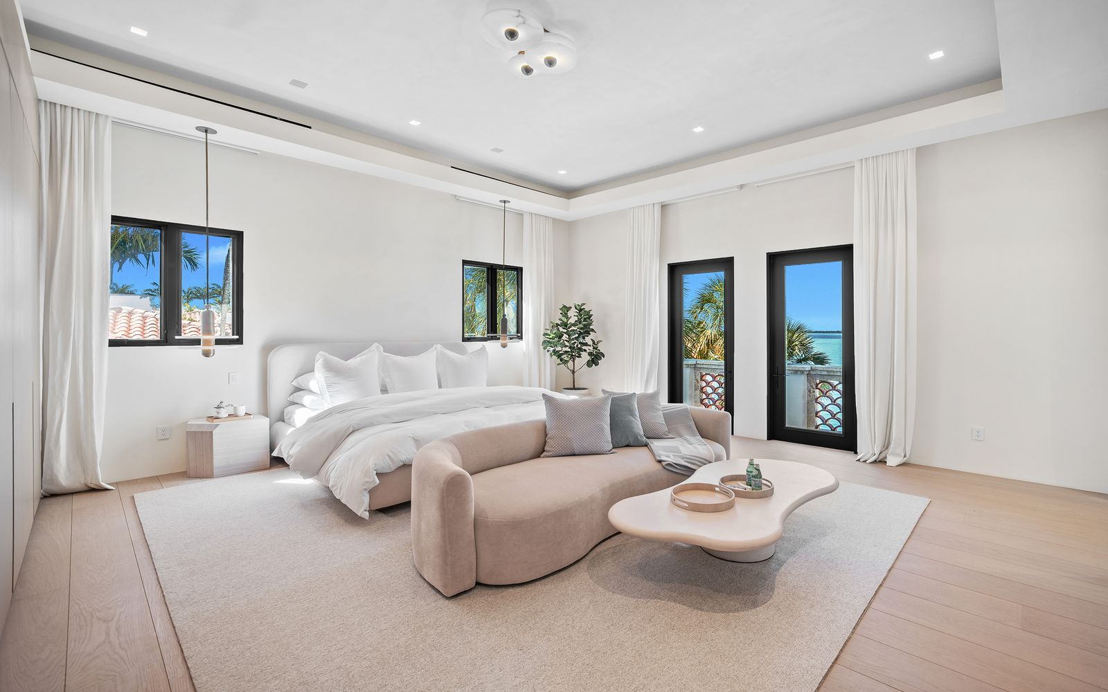 In the bedroom, floor-to-ceiling glass doors open toward a waterfront balcony, while soft neutral finishes, pale wood floors, and minimal lighting fixtures keep the oversized room calm and uncluttered.