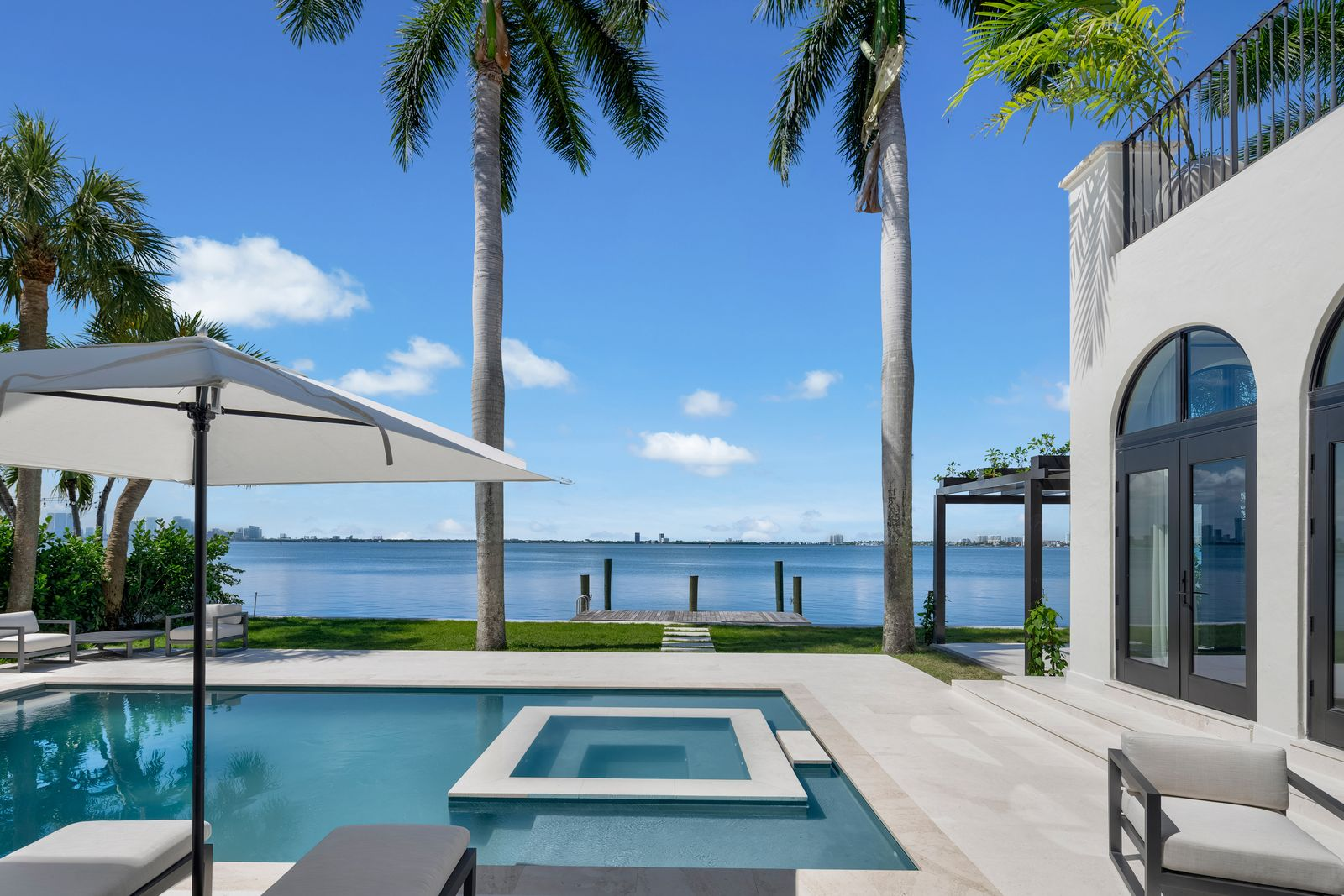 Pale stone decking wraps the outdoor spaces, while rows of palm trees and open sightlines keep the focus on the water and Miami skyline beyond.